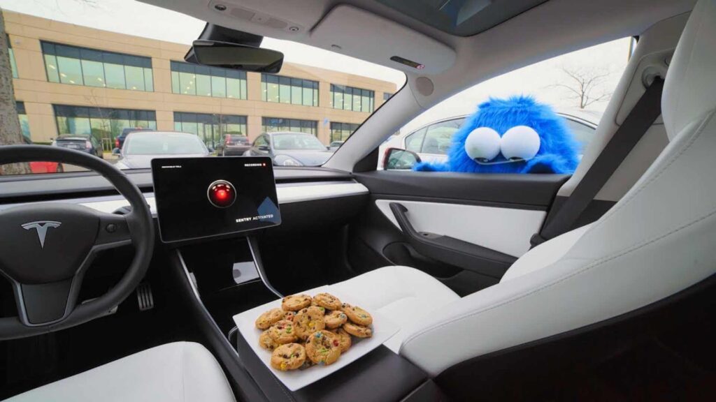 Interiér vozu Tesla s velkou dotykovou obrazovkou, na které je zobrazen text 'Sentry Mode Activated' a červená ikona nahrávání. Na sedadle spolujezdce sedí modrá chlupatá postavička s velkými očima, která připomíná loutku, za níž na středovém konzolu leží talíř s cookies.