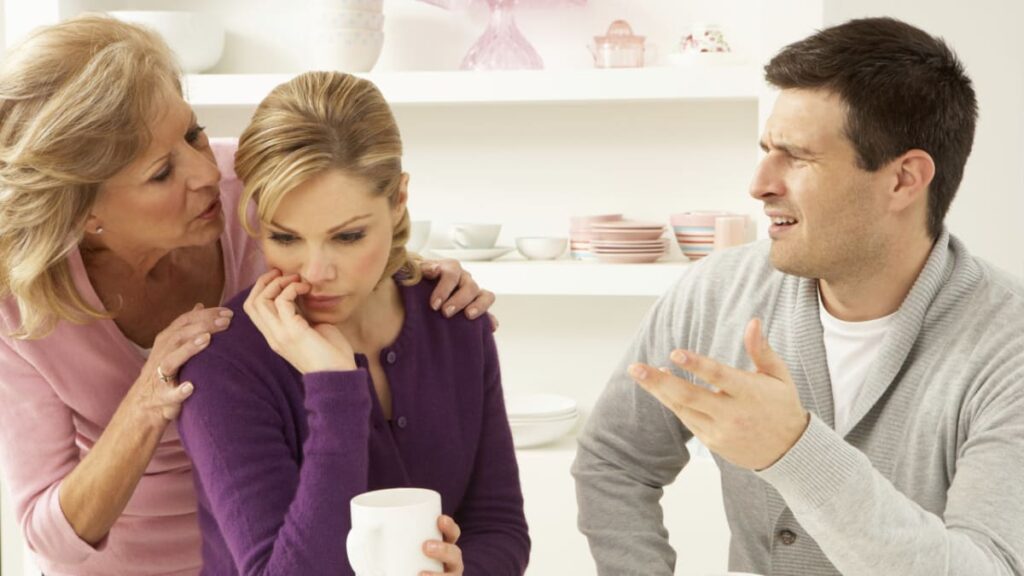 Manželský pár v diskuzi s tchýní, která utěšuje znepokojenou snachu, zatímco manžel gestikuluje v projevu nesouhlasu v domácím prostředí.