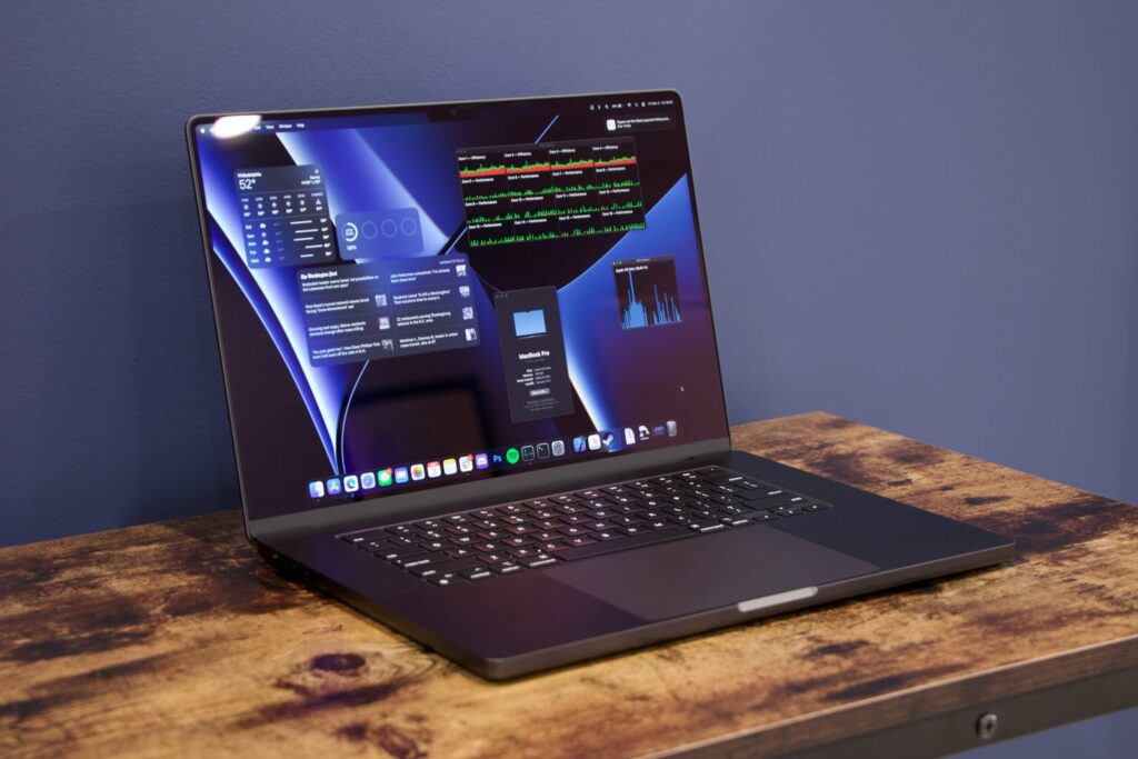 MacBook Pro 16 M3 Max umístěný na dřevěném stole, s otevřeným krytem, zobrazující plochu s aplikacemi na tmavém pozadí s modrým gradientem. Klávesnice je podsvícená a na obrazovce jsou vidět widgety s informacemi o počasí a výkonu počítače.