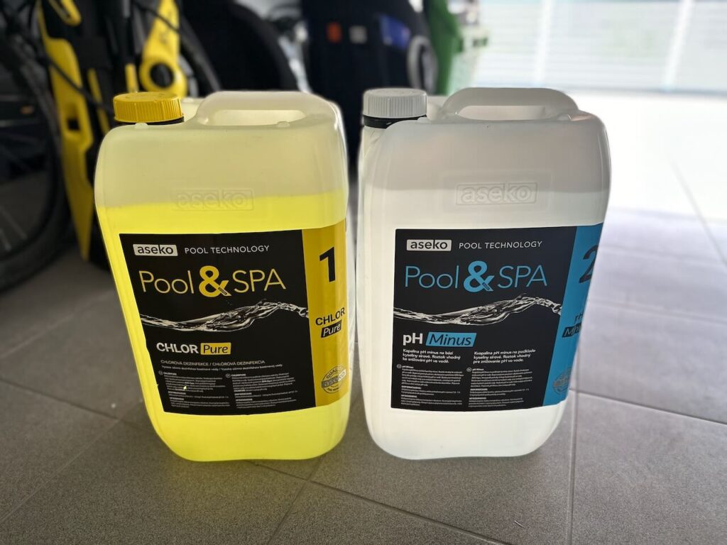 Dva velké plastové kanystry na dlaždicové podlaze v garáži, jeden s nápisem 'Pool & SPA CHLOR Pure' a druhý s nápisem 'Pool & SPA pH Minus', oba od značky Aseko, umístěné před žlutým sportovním autem.