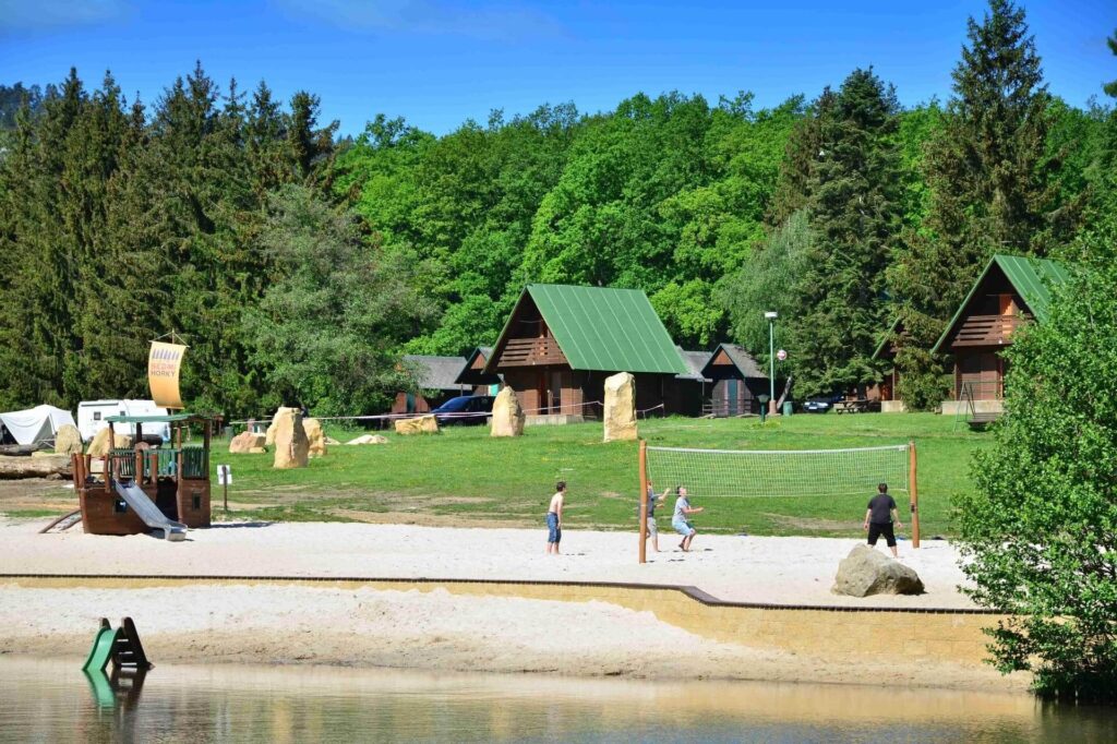 Lidé hrají plážový volejbal v kempu Semihorky s chatkami a dětským hřištěm u jezera.