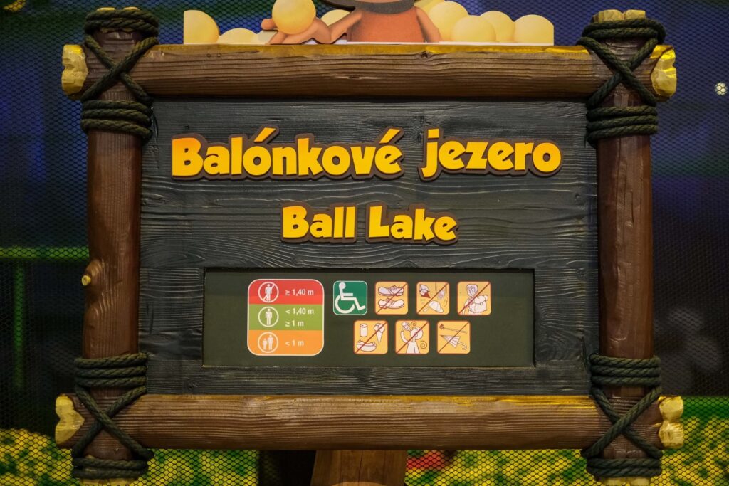 Na obrázku je dřevěný návěstní štít s nápisem "Balónkové jezero" v češtině a pod ním "Ball Lake" v angličtině. Návěstí je umístěno na tmavě hnědém pozadí a je orámováno světle hnědým dřevem s vikingskými ornamenty, podobnými těm, které byly vidět na předchozích fotografiích z parku Majaland. Pod názvy jsou bezpečnostní informace: zelený symbol s "≥ 1,40 m" značí, že návštěvníci této výšky a větší mohou vstoupit sami, zatímco oranžový symbol s "< 1,40 m" signalizuje, že menší děti potřebují dospělý doprovod. Dále jsou zde ikony ukazující bezpečnostní pravidla, jako je například zákaz vstupu s jídlem a pitím, zákaz vstupu těhotným ženám, a podobně. Celý štít má rustikální, tematický design, který odpovídá dětskému hřišti, na kterém se nachází.