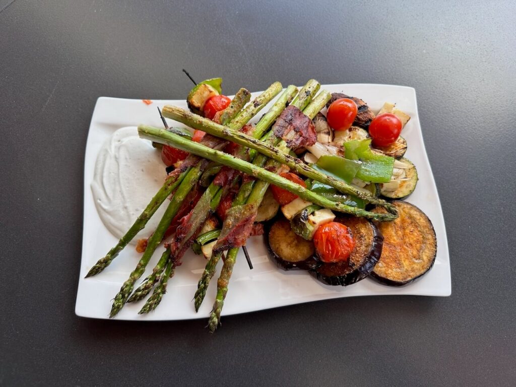 Grilovaná zelenina na bílém talíři s česnekovým dipem – asparagus, cukety, žampiony a cherry rajčátka, doplněné o kousky papriky a obalené v šunce.