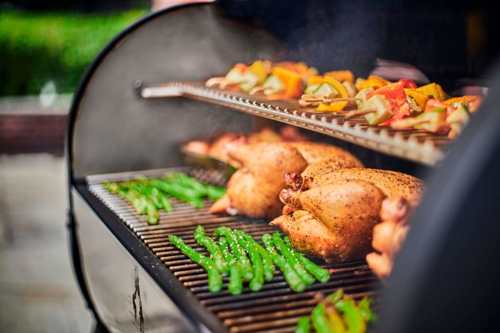 Čerstvé jídlo grilované na Weber grilu, zahrnující kuřata a zeleninové špízy s paprikou a cuketou, spolu se zelenými chřestovými lusky, vše kouřící se nad grilovacími rošty.