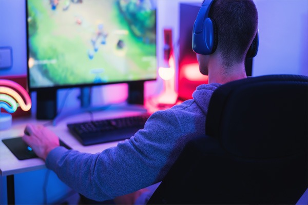 Muž v šedé mikině sedí v herním křesle a hraje videohry na počítači s barevným LED osvětlením.