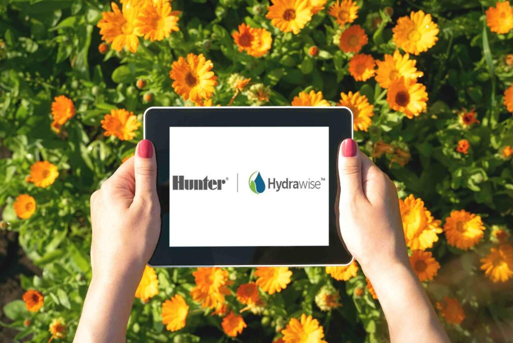 Osoba drží tablet s logem Hunter Hydrawise na obrazovce nad kvetoucí zahrádkou s oranžovými květy, zdůrazňující používání technologie při správě zavlažování.