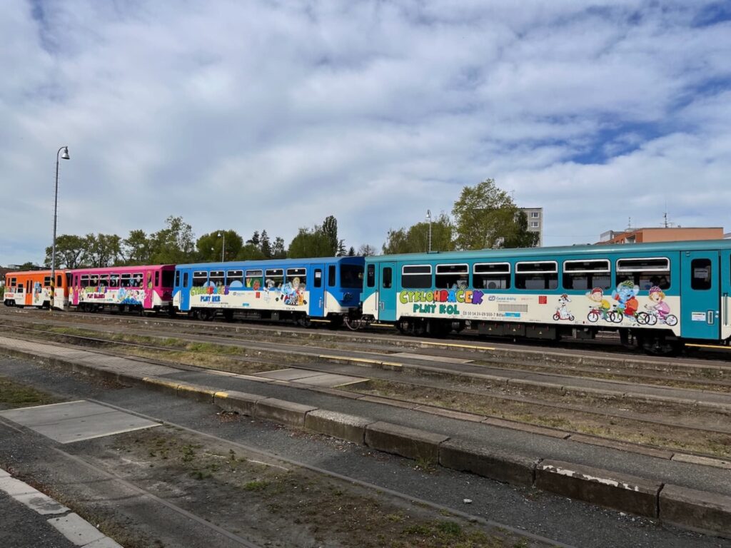 Pestrý vlak Cyklohráček Českých drah stojící na nádraží, s barevnými vagóny a dětskými motivy na boku, pod jasně modrou oblohou.