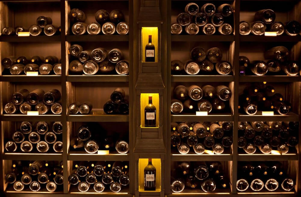 Pohled na tmavě osvětlenou vinnou skříň plnou ležatých lahví vína uspořádaných ve více řadách na dřevěných policích, s vyčnívajícím středovým regálem osvětleným žlutým světlem, kde jsou láhve vystaveny ve výklencích.