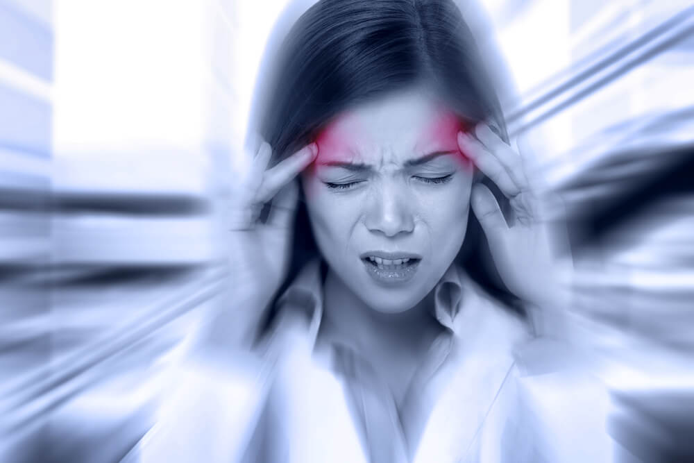 Žena držící si ruce na spáncích s bolestivým výrazem, oblasti bolesti jsou zvýrazněny červenou barvou, kolem je rozmazané pozadí symbolizující závratě nebo silnou bolest hlavy díky migréně.