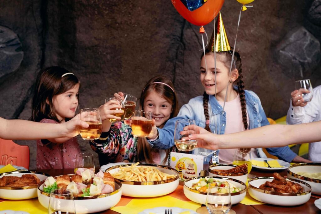 Na fotografii jsou tři děti oslavující narozeniny u stolu s jídlem. Uprostřed sedí děvče s narozeninovým kloboučkem a balónkem, zdá se, že je oslavenkyně. Děti drží sklenice s nápojem a připíjejí si v radostné atmosféře. Na stole je různé jídlo, včetně kuřecích křidýlek, hranolek a sendvičů. Vše je nastaveno pro dětskou oslavu, s jasnými barvami a veselou náladou.