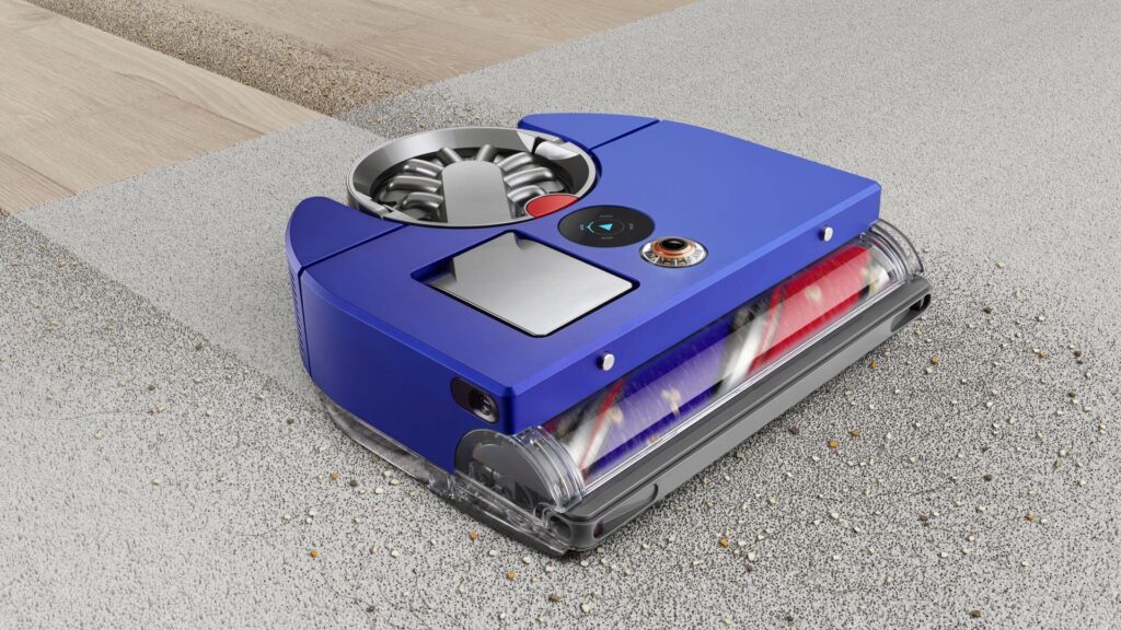 Modrý robotický vysavač Dyson při práci na koberci a tvrdé podlaze, se zametaným odpadem.