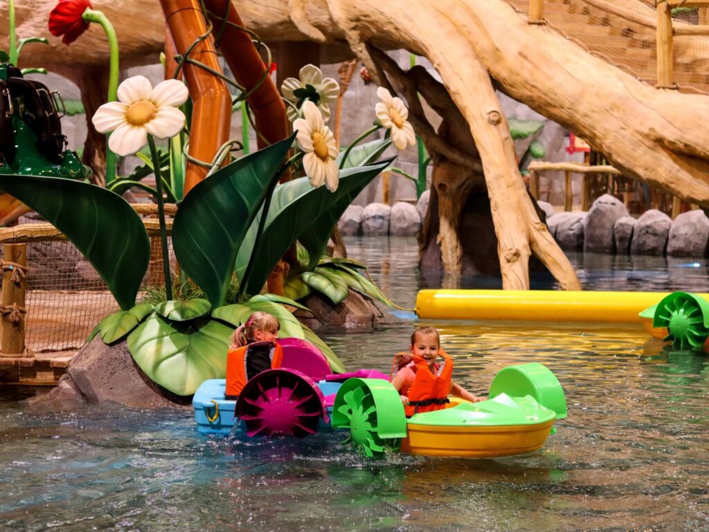 Obrázek zachycuje děti v malých ručních lodičkách na vodní atrakci v interiéru zábavního parku. Lodičky jsou barevné, obklopeny velkými umělými květinami a vegetací, které vytvářejí pocit pohádkového lesa. Děti působí šťastně, jak se plaví po vodní hladině, a atmosféra je hravá a bezpečná s měkkými, zaoblenými tvary a výraznými barvami.