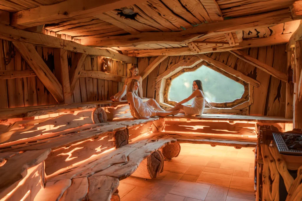 Tři ženy v relaxační póze ve dřevěné sauně, s teplým světlem pronikajícím skrz jedinečné, asymetrické okno. Sauna má organický design, s nepravidelně tvarovanými trámy a hladce opracovanými dřevěnými lavicemi, které vyzařují přírodní a uklidňující atmosféru. Místo poskytuje pocit soukromí a klidu, ideální pro odpočinek a regeneraci.