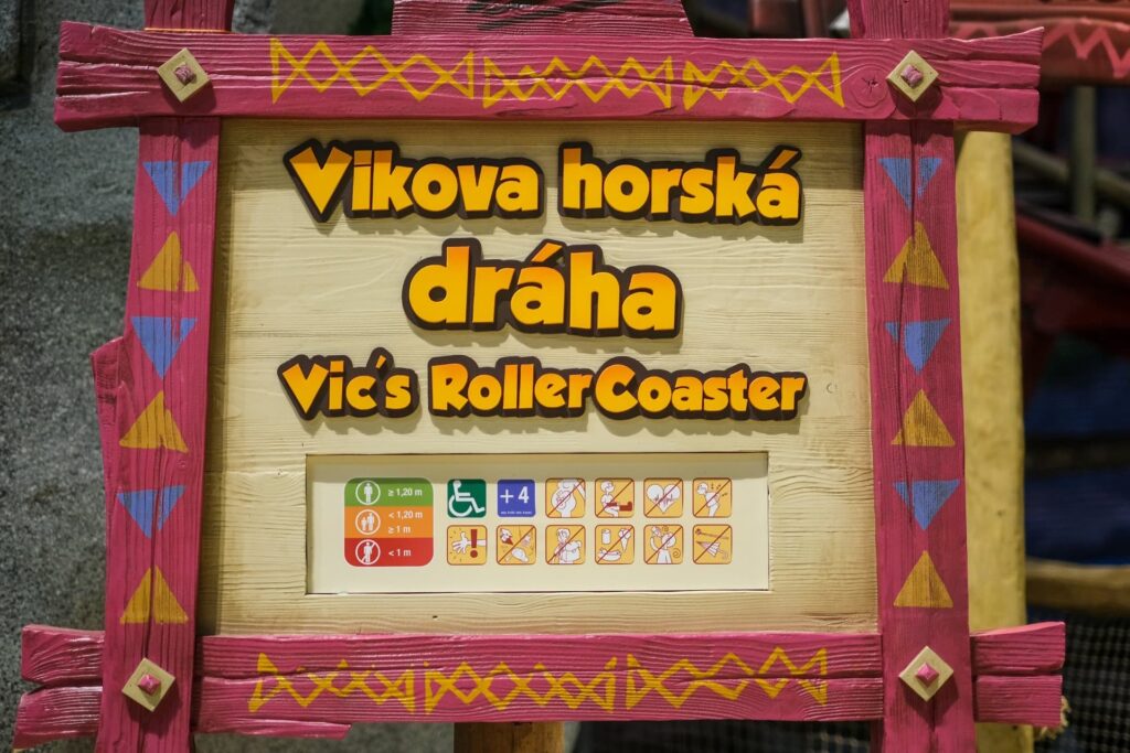 Na fotografii je tabule s nápisem "Vikova horská dráha" a pod tím anglickým překladem "Vic's Roller Coaster". Rámeček tabule je dřevěný s ružovými a žlutými vikingskými motivy. Pod nápisy jsou bezpečnostní a výškové informace pro návštěvníky atrakce: zelený kroužek s "+1.20 m" značí, že lidé vyšší než 1,20 metru mohou jít sami, oranžová ikona s "-1.20 m" značí, že ti menší než 1,20 metru musí být doprovázeni dospělým. Dále jsou tu ikony s bezpečnostními pravidly, jako je zákaz vstupu těhotným ženám, lidem se zdravotními problémy srdce atd. Atmosféra je hravá a dekorace podtrhují tematiku atrakce.