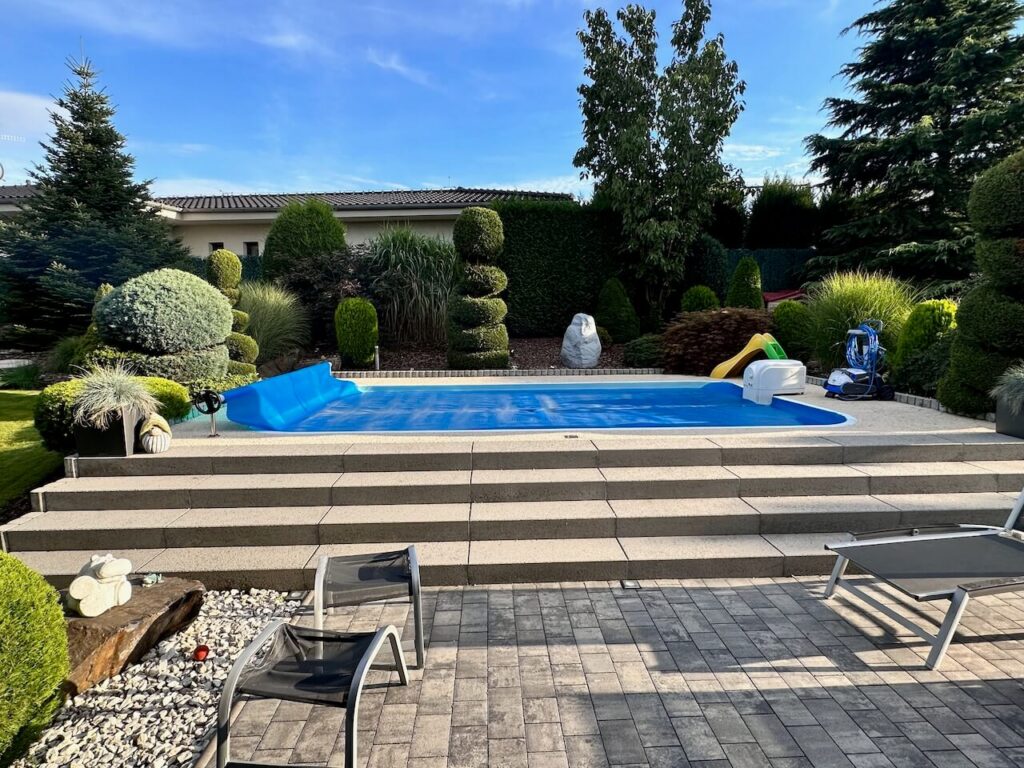 Zahrada s bazénem s modrou vodou obklopený upravenou zahradou s různými stromy a keři, kamennými chodníčky a dvěma lehátky, s hřištěm a robotickým vysavačem pro bazén na pravé straně.