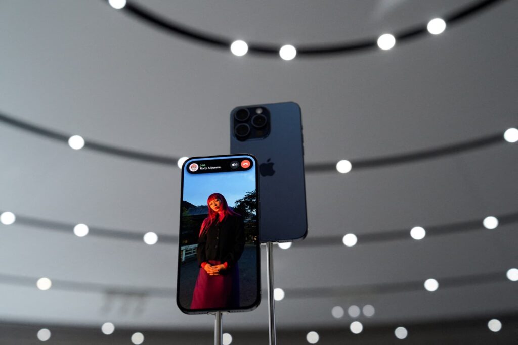 Apple iPhone 15 pro max na stojanu zobrazuje ženu s rudými vlasy a v červeném oblečení, která stojí venku za soumraku. V pozadí je vidět venkovskou scenérii s domem. Telefon je před abstraktním pozadím s kruhovými světly.