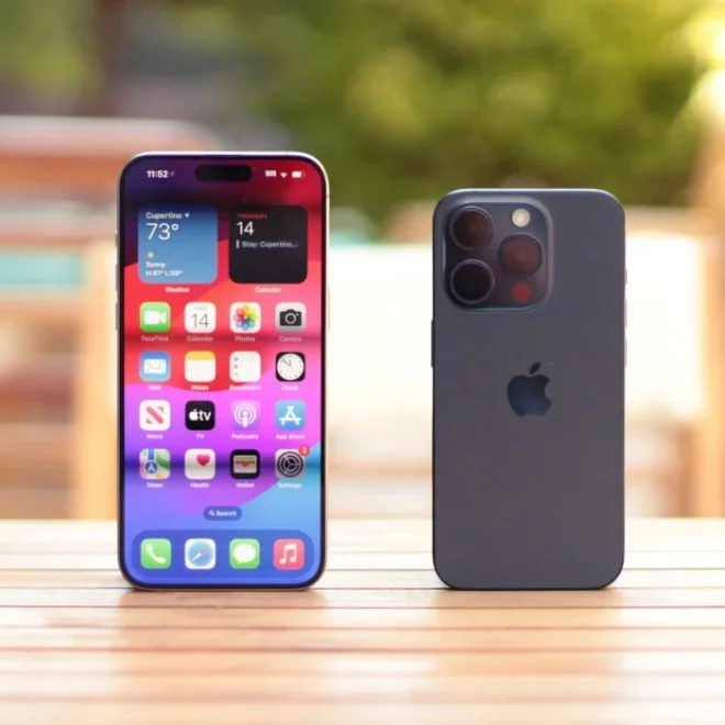 Dva iPhone modely jsou postaveny na dřevěném stole venku. Na levém telefonu je rozsvícený displej ukazující plochu s ikonami aplikací a informacemi o počasí v Cupertino. Pravý telefon je otočený zadní stranou s viditelnými fotoaparáty, umístěný na pozadí s rozostřenou zahradou a slunečním svitem.