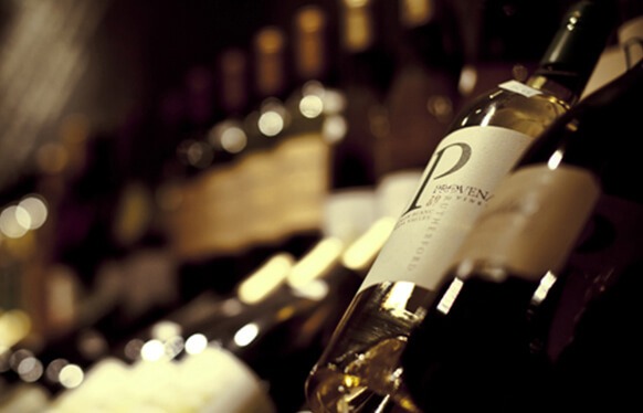 Rozostřený pohled na řadu ležících lahví vína s detailně zobrazenou etiketou na přední lahvi, v atmosféře zlatavého osvětlení typického pro vinotéku.