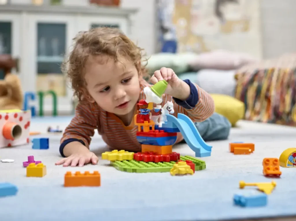 Dítě se soustředěným výrazem hraje na koberci s LEGO DUPLO sady, skládá postavičky a klouzačku, obklopené rozptýlenými barevnými kostkami.