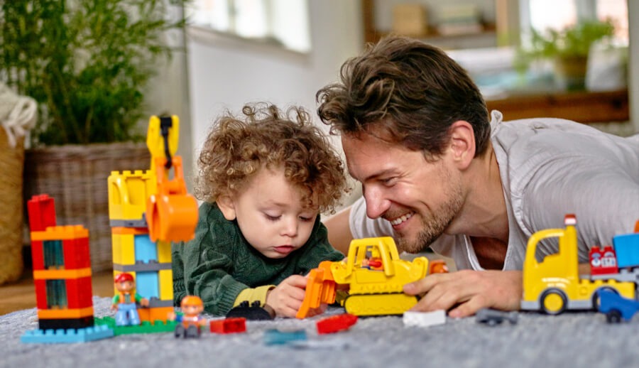 Otec a malý chlapec hrající si na podlaze s LEGO DUPLO stavebnicemi, včetně žlutého bagru a nákladního auta, stavějí barevné konstrukce.