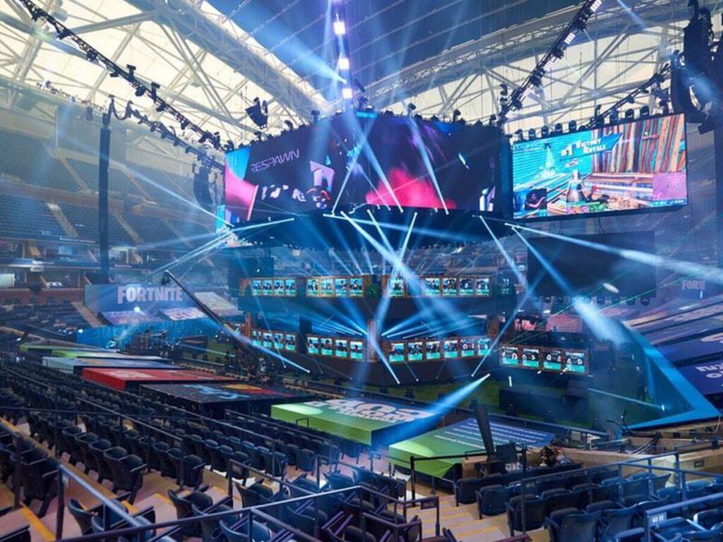 Pohled na profesionální esportovní arénu Fortnite před zápasem, osvětlenou modrými reflektory a s velkými obrazovkami zobrazujícími hru.