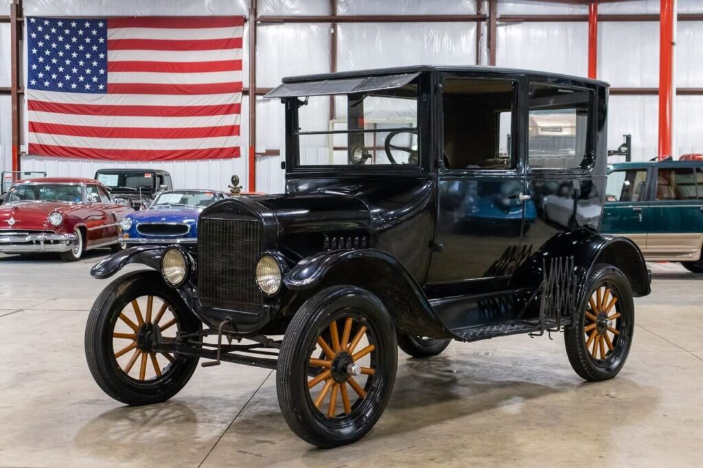 Černý Ford Model T z roku 1925 vystavený v automobilovém muzeu před americkou vlajkou. Tento historický vůz je symbolem revoluce v automobilovém průmyslu, která přinesla cenově dostupná auta pro širokou veřejnost. V pozadí jsou vidět další klasické vozy.