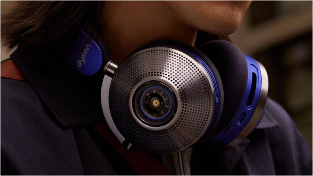 Detailní záběr na moderní sluchátka Dyson se šedými kovovými mřížkami a modrými prvky, nošená mužem v tmavém obleku. Sluchátka mají výrazný design a jsou zaměřená na kvalitu zvuku a pohodlí.