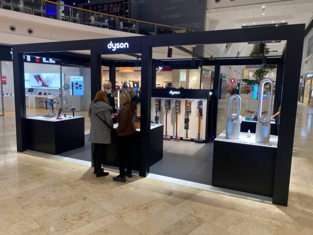 Stánek značky Dyson v nákupním centru s prezentací výrobků, včetně vysavačů a ventilátorů, s třemi zákazníky, kteří se dívají na produkty.