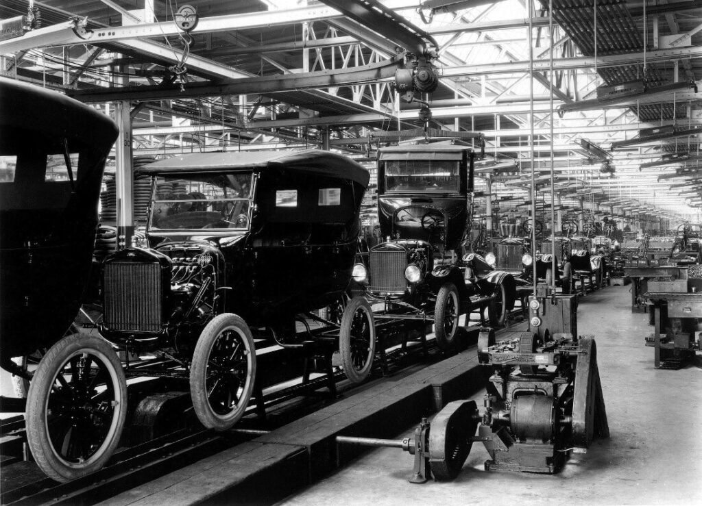 Černobílá fotografie továrny Ford z počátku 20. století, kde jsou na montážní lince sestavovány automobily Ford Model T. Tento revoluční výrobní proces umožnil masovou produkci automobilů, což výrazně snížilo jejich cenu a zpřístupnilo je široké veřejnosti.