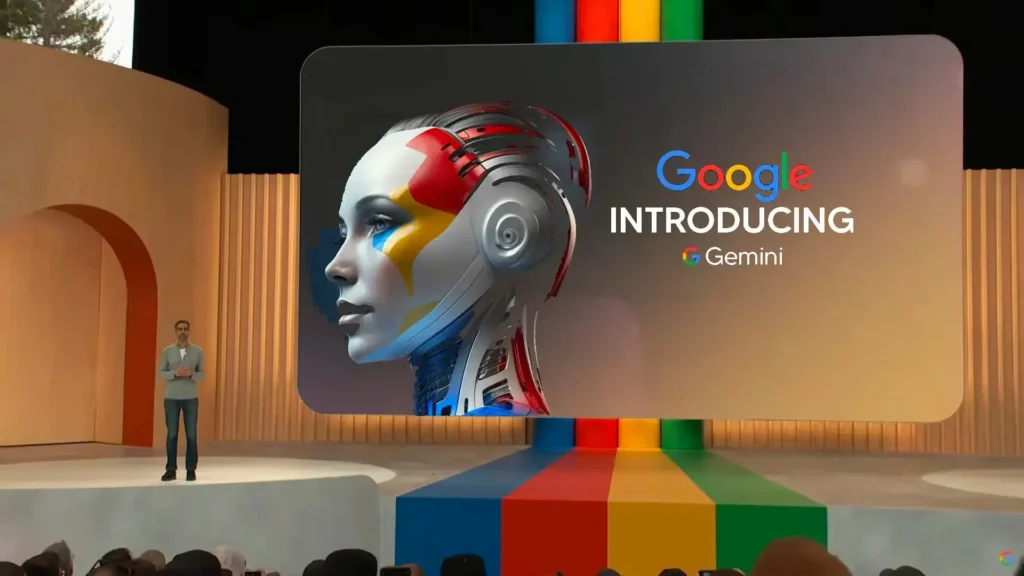 Na obrázku je prezentace Google Gemini, kde na pódiu stojí Sundar Pichai, generální ředitel Google, před velkou obrazovkou. Na obrazovce je zobrazen futuristický portrét humanoidního robota s textem "Google Introducing Gemini". Scéna je moderně navržená s pestrobarevným kobercem a minimalistickým designem. Prezentace představuje nový AI systém Google Gemini.