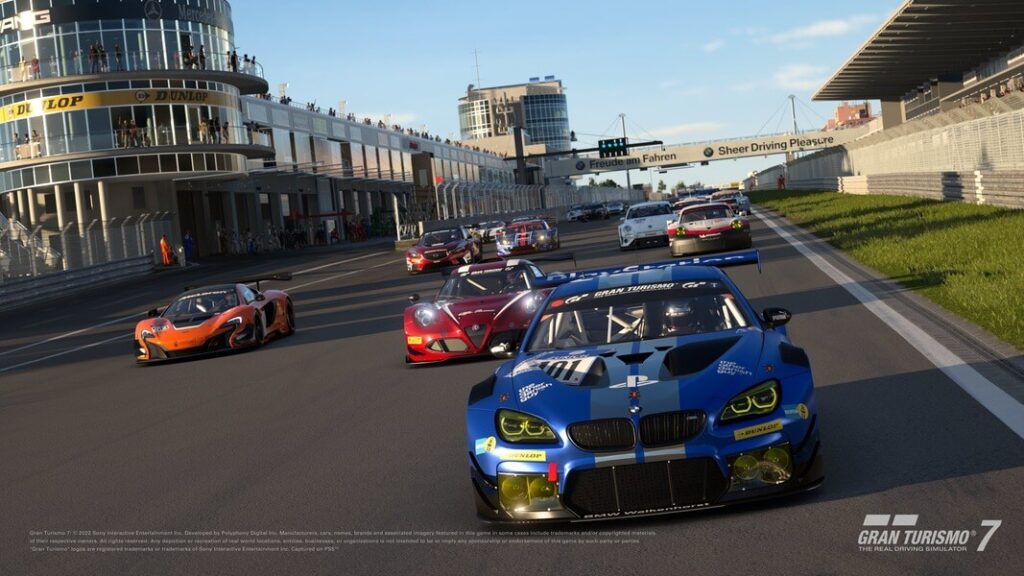 Závodní auta stojí připravena na startovní čáře, v popředí je modré BMW GT s dalšími vozy za ním, včetně konceptních modelů Porsche a Mazda, v simulaci závodů ve hře Gran Turismo 7.