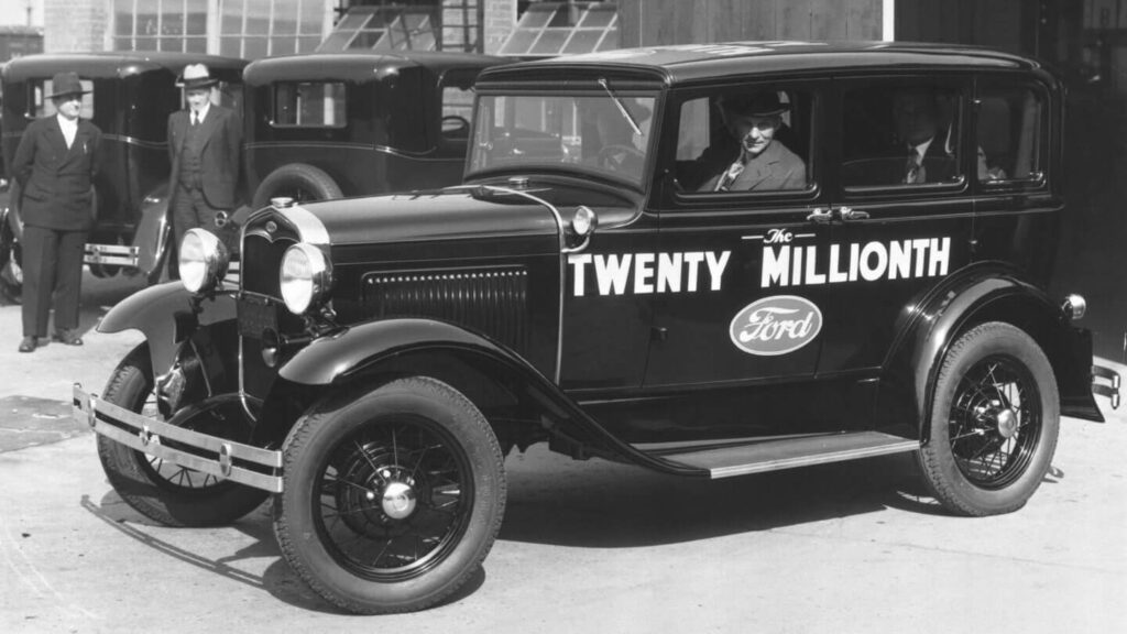 Černobílá fotografie zobrazuje Henryho Forda sedícího v automobilu označeném jako "Twenty Millionth Ford", což znamená dvacátý miliontý vyrobený vůz Ford. Vedle auta stojí několik mužů v kloboucích a oblecích. Fotografie zachycuje významný milník v historii automobilky Ford.