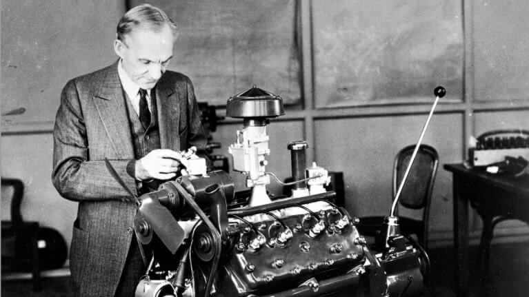 Henry Ford pracuje na motoru v dílně. Je oblečený v obleku, soustředěně se věnuje technickému detailu motoru, který má před sebou. Tato fotografie zachycuje Fordovu vášeň pro techniku a jeho praktický přístup k inženýrství a inovacím.