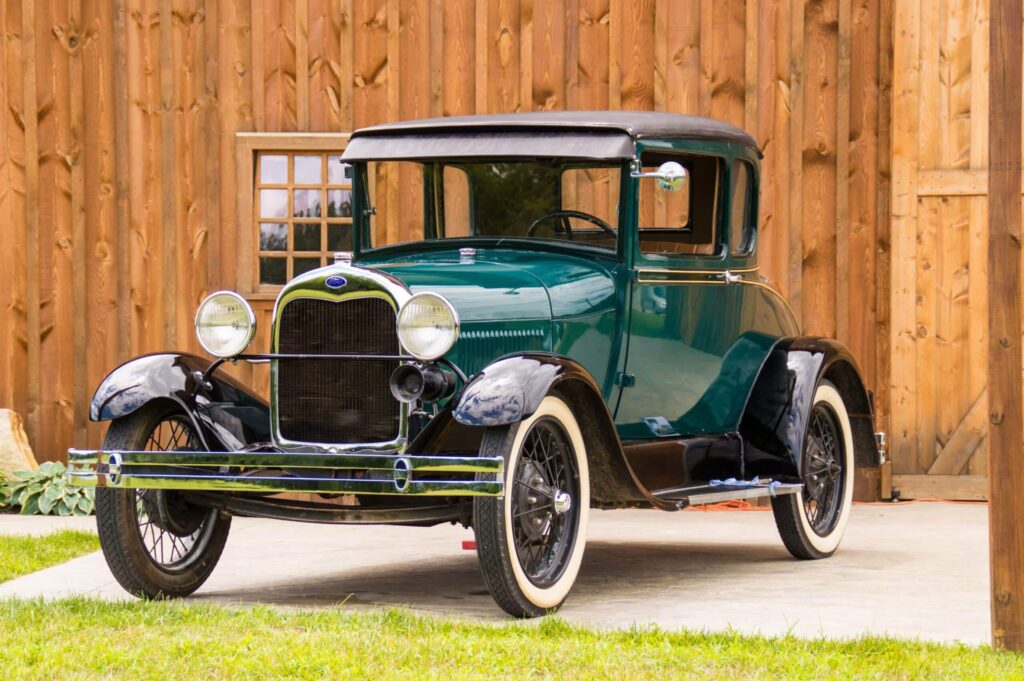 Elegantní zelený Ford Model T z roku 1925 s černými blatníky a bílými pneumatikami, zaparkovaný před dřevěnou budovou. Tento historický vůz je ikonou automobilového průmyslu a představuje technologický pokrok své doby.