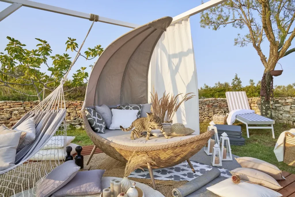 Moderní zahradní sezení s lehátkem ve tvaru skořápky, doplněné houpací sítí a pohodlnými polštáři, situované v malebné zahradě. Na lehátku leží kočka, okolní prostor je dekorován přírodními prvky a dekorativními lampami.