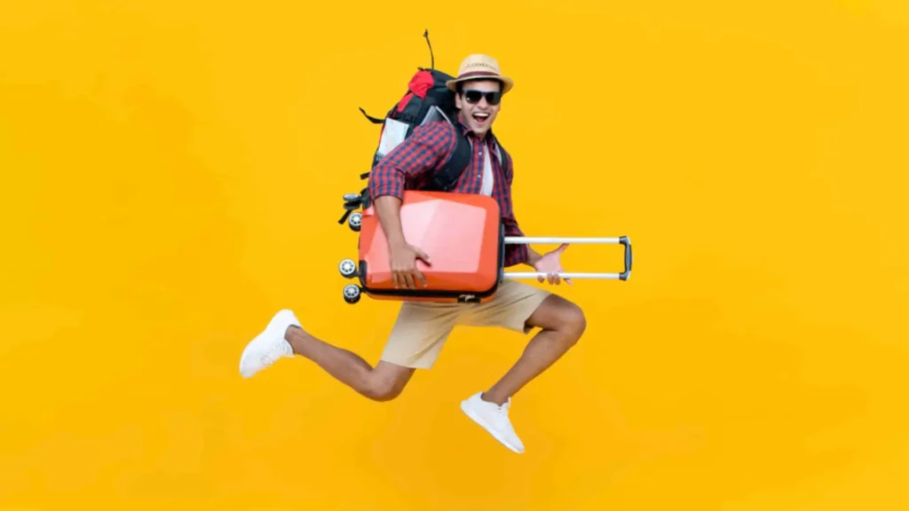 Šťastný mladý muž v klobouku, slunečních brýlích a s batohem na zádech drží oranžový kufr a skáče radostí na žlutém pozadí.
