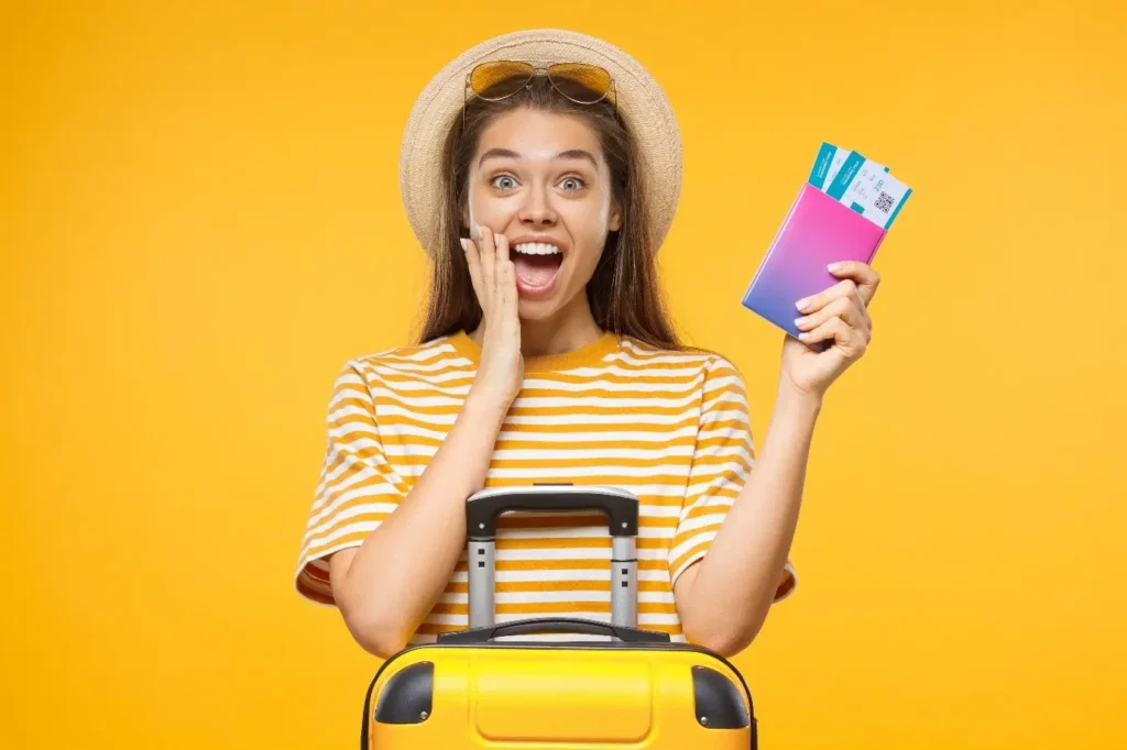 Šťastná mladá žena v pruhovaném tričku a slamáku drží v ruce cestovní pas s letenkami, stojící vedle žlutého kufru na kolečkách, na žlutém pozadí.