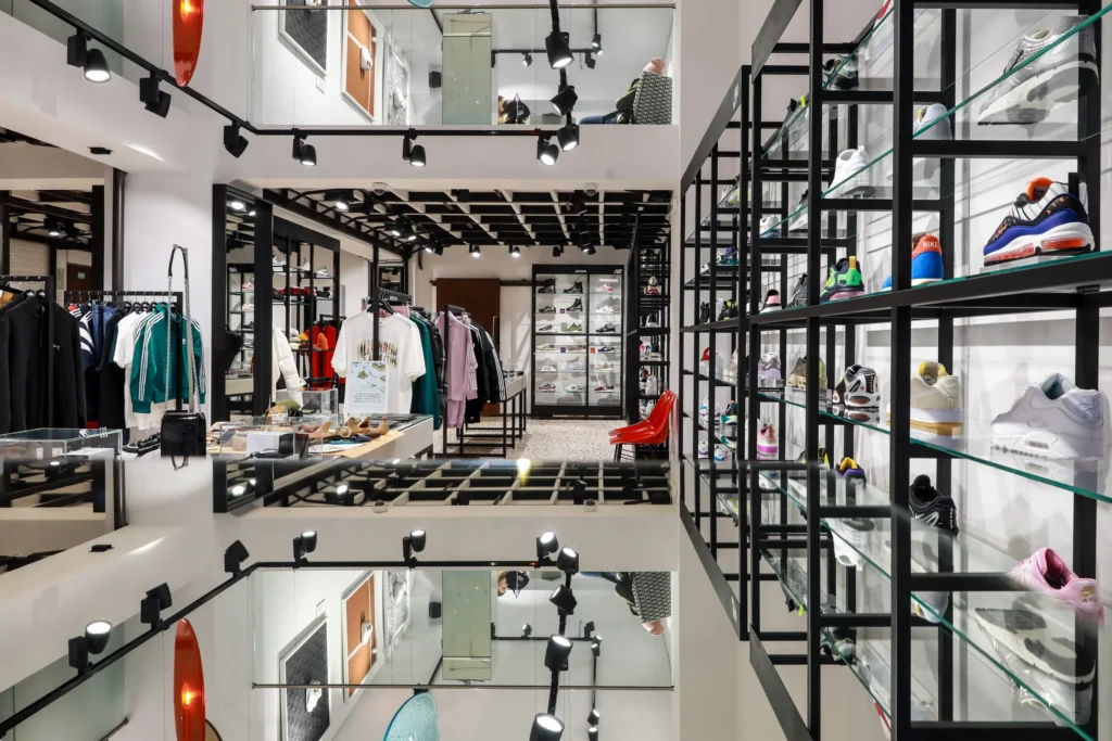 Interiér prodejny Footshop s vystavenými teniskami a streetwearovým oblečením. Na policích jsou různobarevné tenisky a stojany s oblečením. Moderní design obchodu zahrnuje zrcadla a stylové osvětlení.