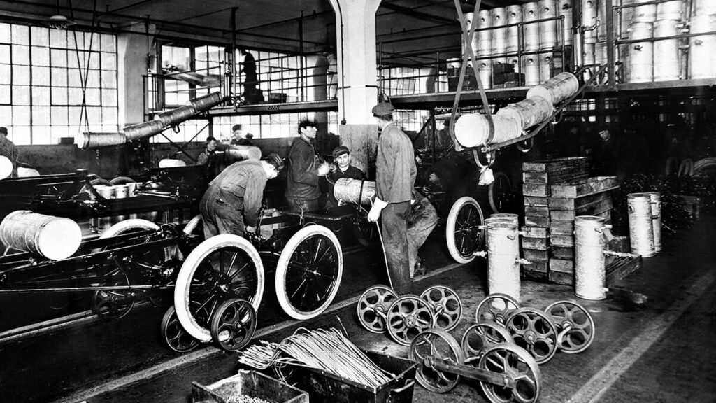 Historická fotografie z továrny Ford Motor Company ukazuje dělníky pracující na montážní lince. Několik mužů sestavuje automobilové šasi a další komponenty za pomoci velkých válcových součástek zavěšených na jeřábu. Továrna je plná dílenského vybavení a ukazuje inovativní montážní proces zavedený Henrym Fordem.