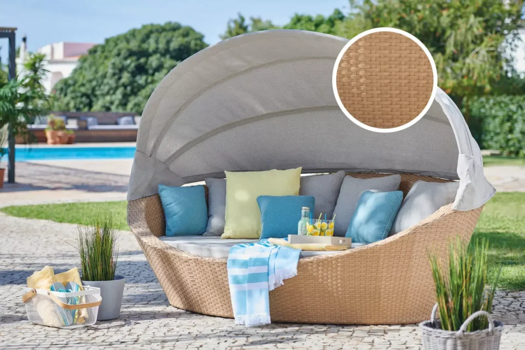Elegantní ratanové lehátko s baldachýnem v moderním designu, obklopené polštáři v pastelových barvách, umístěné na dlážděné ploše u bazénu. Vedle lehátka jsou dekorativní prvky jako rostliny v květináčích a koš s plážovým vybavením.