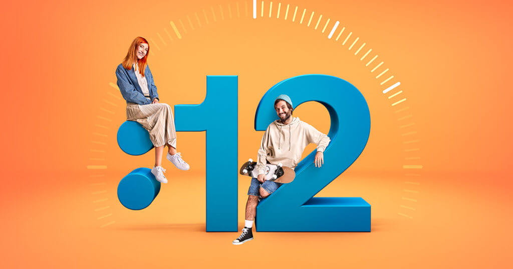 Kreativní reklamní obrázek Razdva půjčka s oranžovým pozadím zobrazující dvě osoby sedící na obrovském modrém čísle 12. Žena s dlouhými rudými vlasy sedí na horní části čísla '1', oblečená v modré džínové bundě a béžových kalhotách. Muž s tmavou vousatou tváří a v bílém svetru sedí na dolní křivce čísla '2', obklopen stylizovanými bílými čárami, které vyjadřují rychlost a dynamiku.