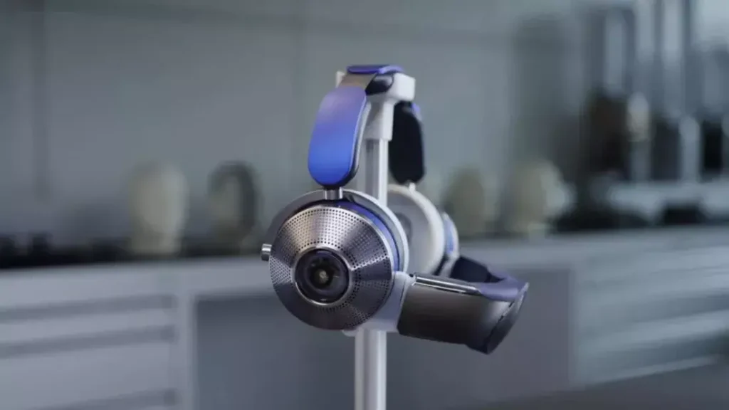 Moderní šedé sluchátka Dyson Zone s modrými detaily, zavěšené na stojanu, s pozadím kuchyňské linky a dalších domácích spotřebičů.
