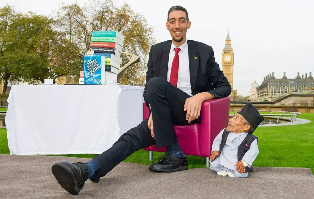 Sultan Kösen, nejvyšší muž světa, sedí vedle nejmenšího muže na světě na křesle v parku s výhledem na Big Ben, vedle nich stojí stůl s knihami Guinnessovy knihy rekordů