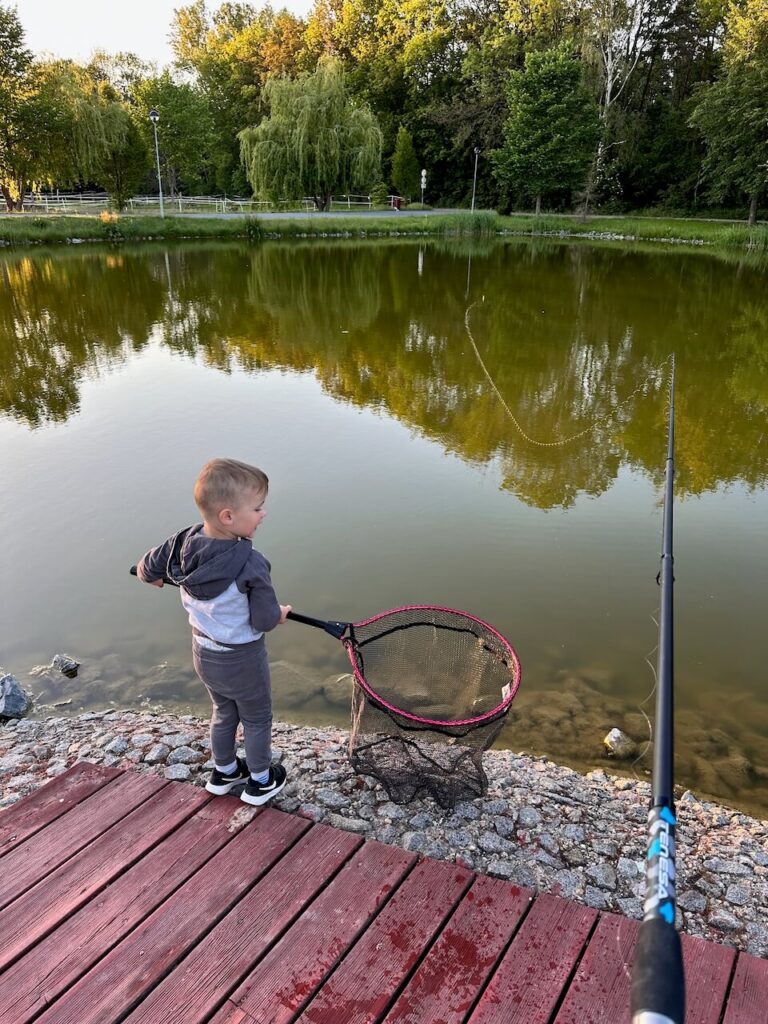 Malý chlapec drží rybářský podběrák na molu u rybníka Amerika v Radonicích, připravený chytit rybu, zatímco rybářský prut čeká na záběr.