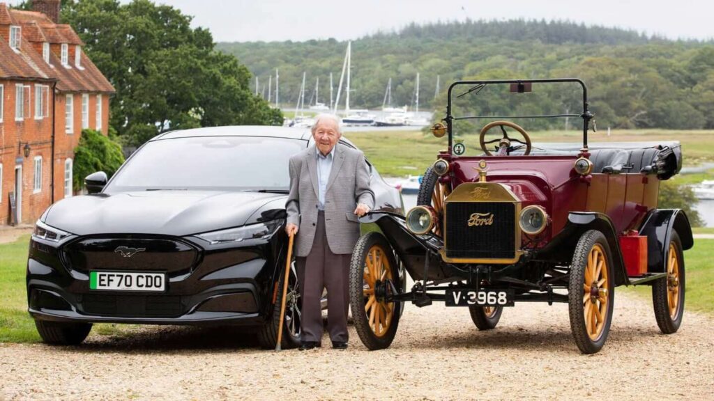 Starší muž stojící mezi dvěma generacemi automobilů Ford: vlevo moderní černý Ford Mustang Mach-E a vpravo historický červený Ford Model T. Fotografie zachycuje kontrast mezi revolučními inovacemi minulosti a současnosti, ilustrující pokrok v automobilovém průmyslu.