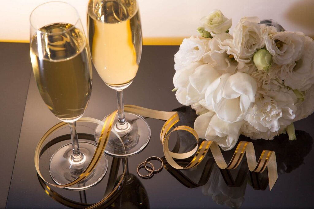 Dva sklenice šampaňského na zrcadlovém podnosu s bílou svatební kyticí a zlatými prstýnky, zdobené zlatou stužkou.