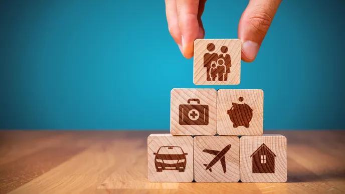 Ruka umísťuje dřevěnou kostku s ikonou rodiny na vrchol pyramidy z dřevěných kostek, na kterých jsou zobrazeny ikony zdravotní péče, ochrany, automobilu, cestování a domova, symbolizující různé typy pojištění.