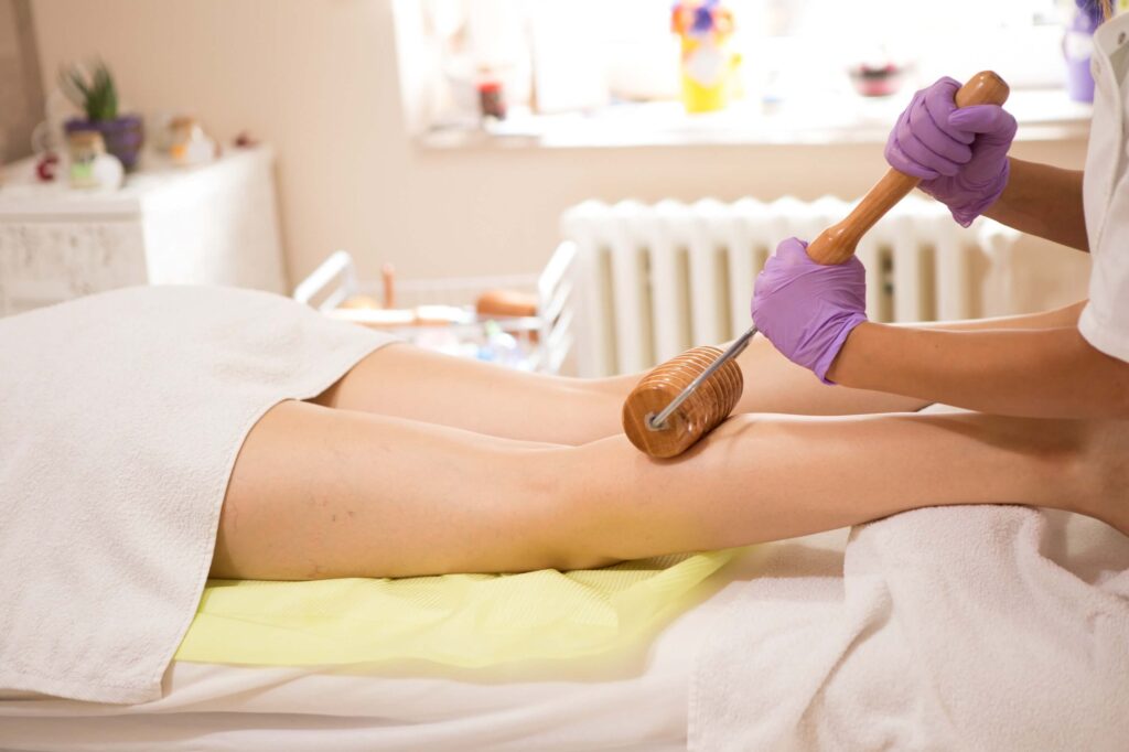 Profesionální masáž proti celulitidě pomocí dřevěného válečku, prováděná v salonu s fialově oblečenou masérkou, která aplikuje masáž na stehna klientky ležící na masážním stole.