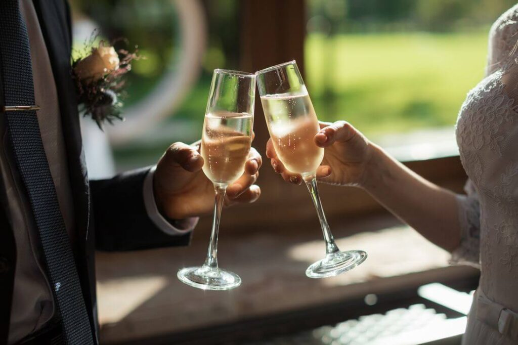 Novomanželé si přiťukávají sklenicemi se šampaňským během svatební oslavy, v pozadí světlý výhled do zeleně.