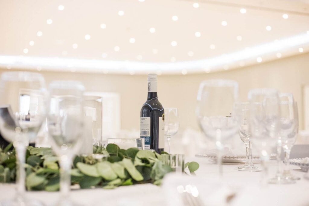 Svatební stůl s lahvi vína uprostřed, obklopený elegantně uspořádanými sklenicemi a zelenými dekoracemi, s jemnými světly v pozadí.